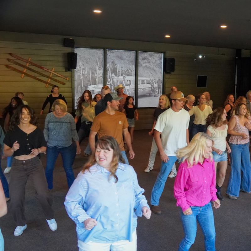 A group of people line dancing in an open room. The atmosphere is lively with smiling faces and a sense of motion, creating an exciting, spirited vibe.