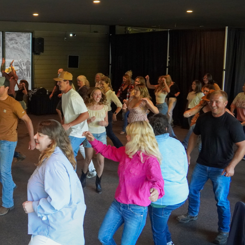 A group of people line dancing in an open room. The atmosphere is lively with smiling faces and a sense of motion, creating an exciting, spirited vibe.