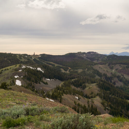 A view of forested mountains with a couple snow patches remaining. The sky has a mixture of lighter and darker clouds, creating a moody atmosphere