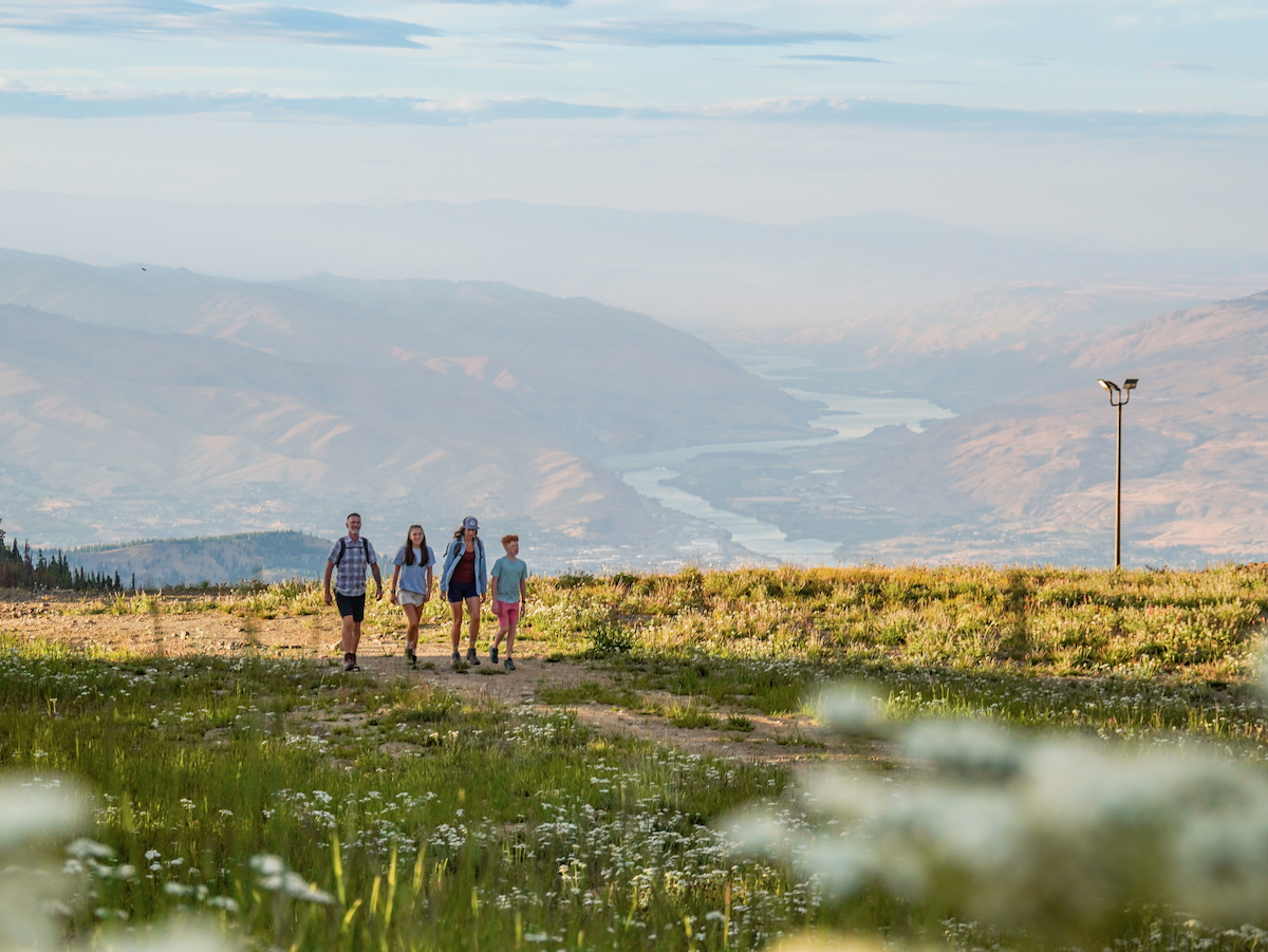 A family walks up a grass and flower-lined path with a view of the Wenatchee Valley and Columbia River in the background far below.