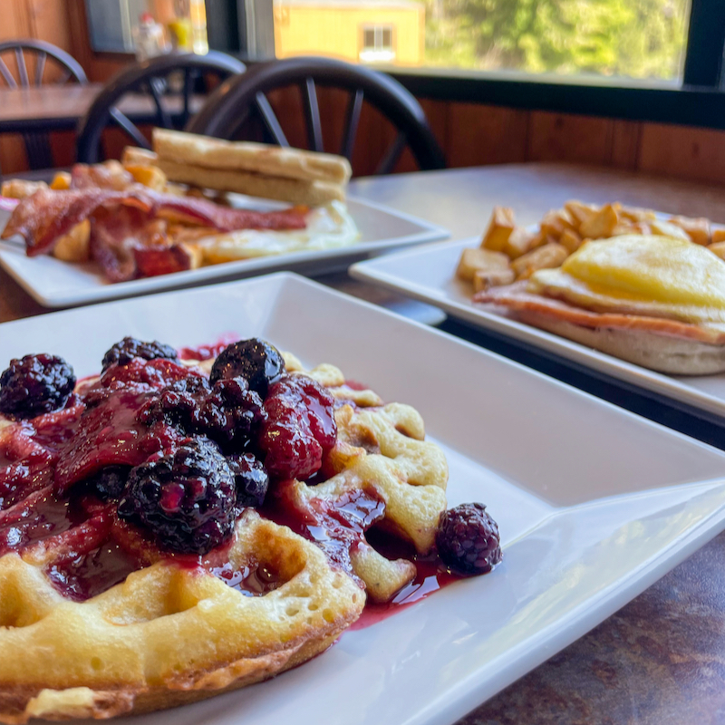 A close-up of a golden waffle topped with mixed berries and syrup on a white plate, with plates of breakfast food including bacon, eggs, toast, potatoes, and pancakes blurred in the background, set on a wooden table near a sunny window.