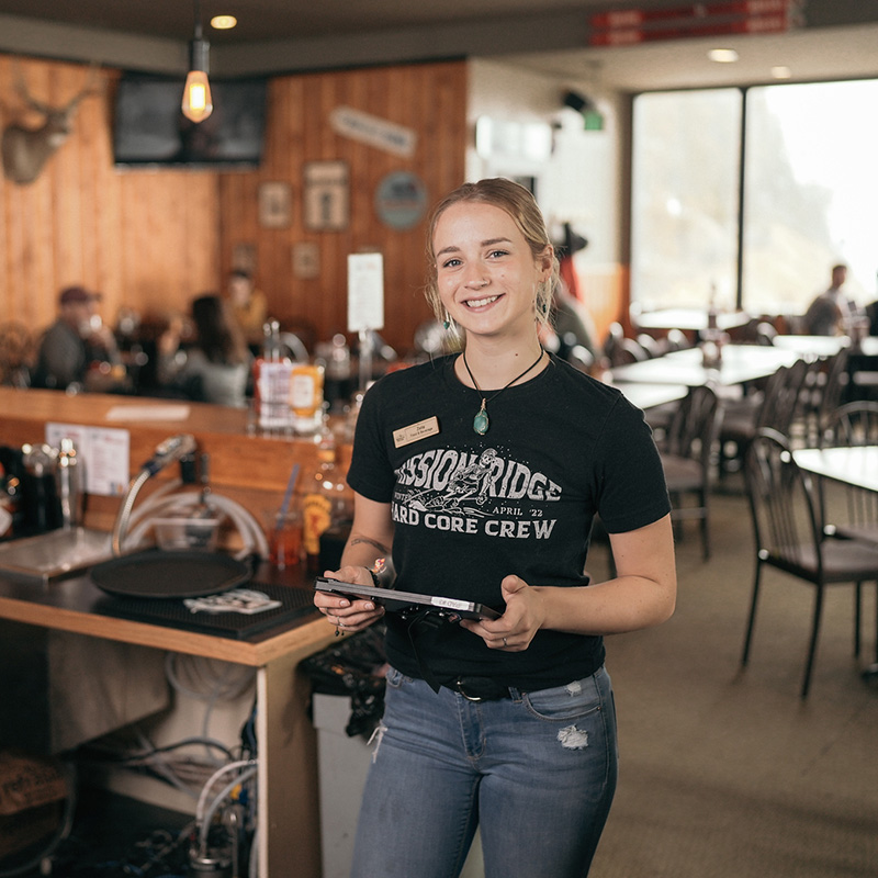 Smiling server wearing a “Mission Ridge Hard Core Crew” t-shirt stands in a cozy mountain restaurant, holding a tablet with tables and customers in the background.