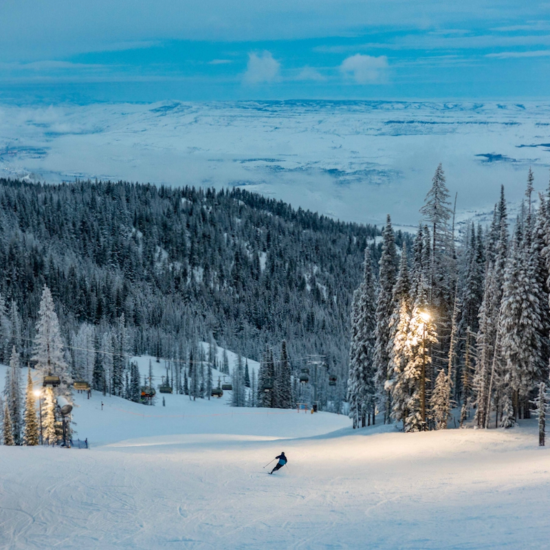 A skier glides down a wide, snow-covered slope at dusk, surrounded by tall, snow-draped evergreen trees. Ski lift chairs hang in the background, while warm lights illuminate the slope, contrasting with the cool blue tones of the snowy mountain landscape and cloudy sky beyond.