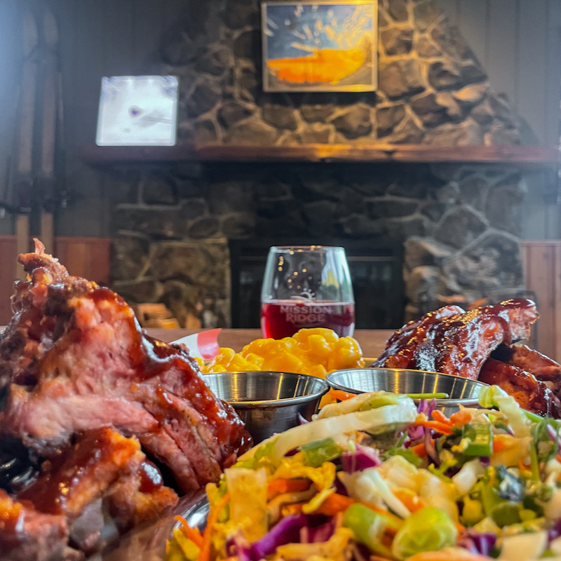A full platter of ribs, coleslaw, and Mac n cheese from the chair 5 Pub at Mission Ridge, with a cozy fireplace creating a fall ambiance in the background.