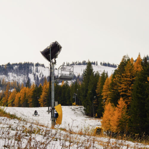 Snowmaking equipment stands on a lightly snow-covered ski slope surrounded by evergreen and golden autumn trees, with mountain ridges in the background.