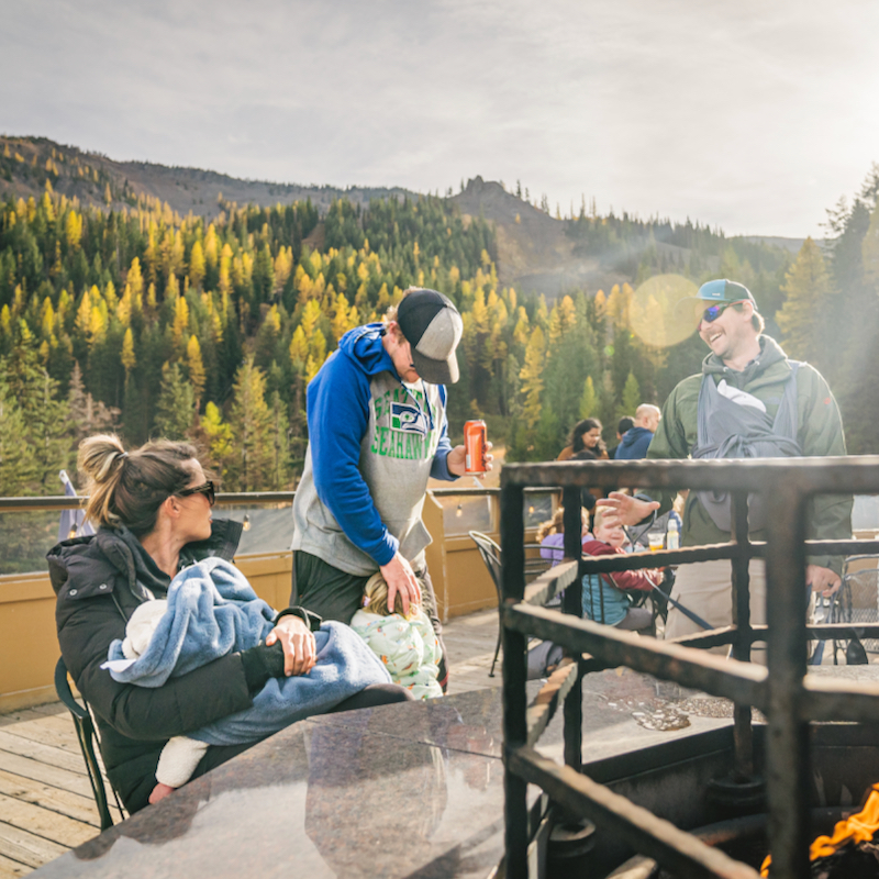 A group of people smiles and laughs on the lodge deck at Mission Ridge, with fall tamaracks painting the hillside gold in the background.