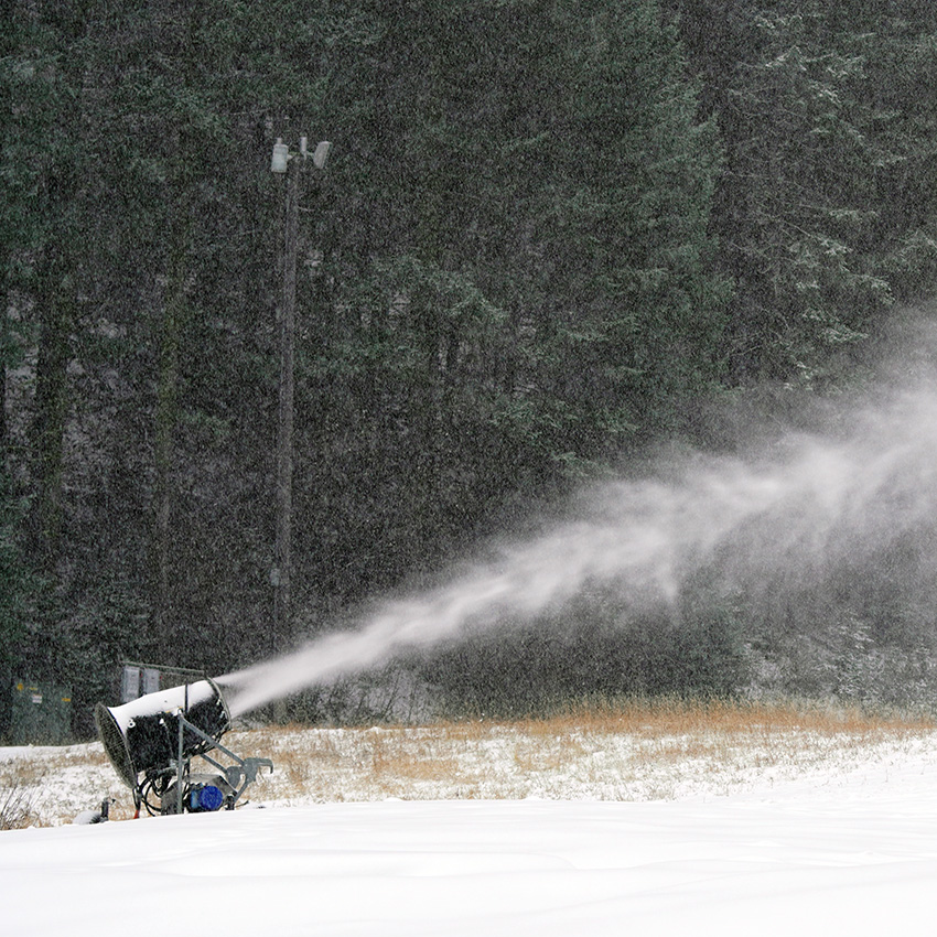 A close-up of a snow gun blasting a plume of snow across a grassy base area, with snowflakes falling and evergreen trees in the background.