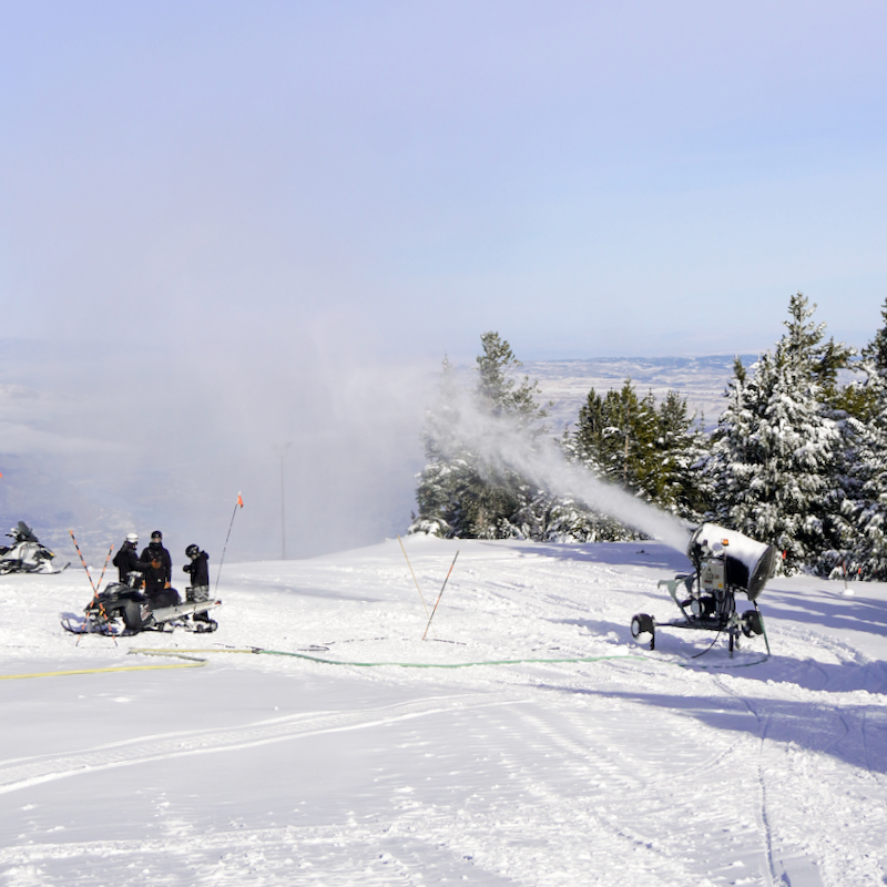 sunny day at the top of Sunspot trail at Mission Ridge, with a snow gun firing and a team of 3 crew members talking.