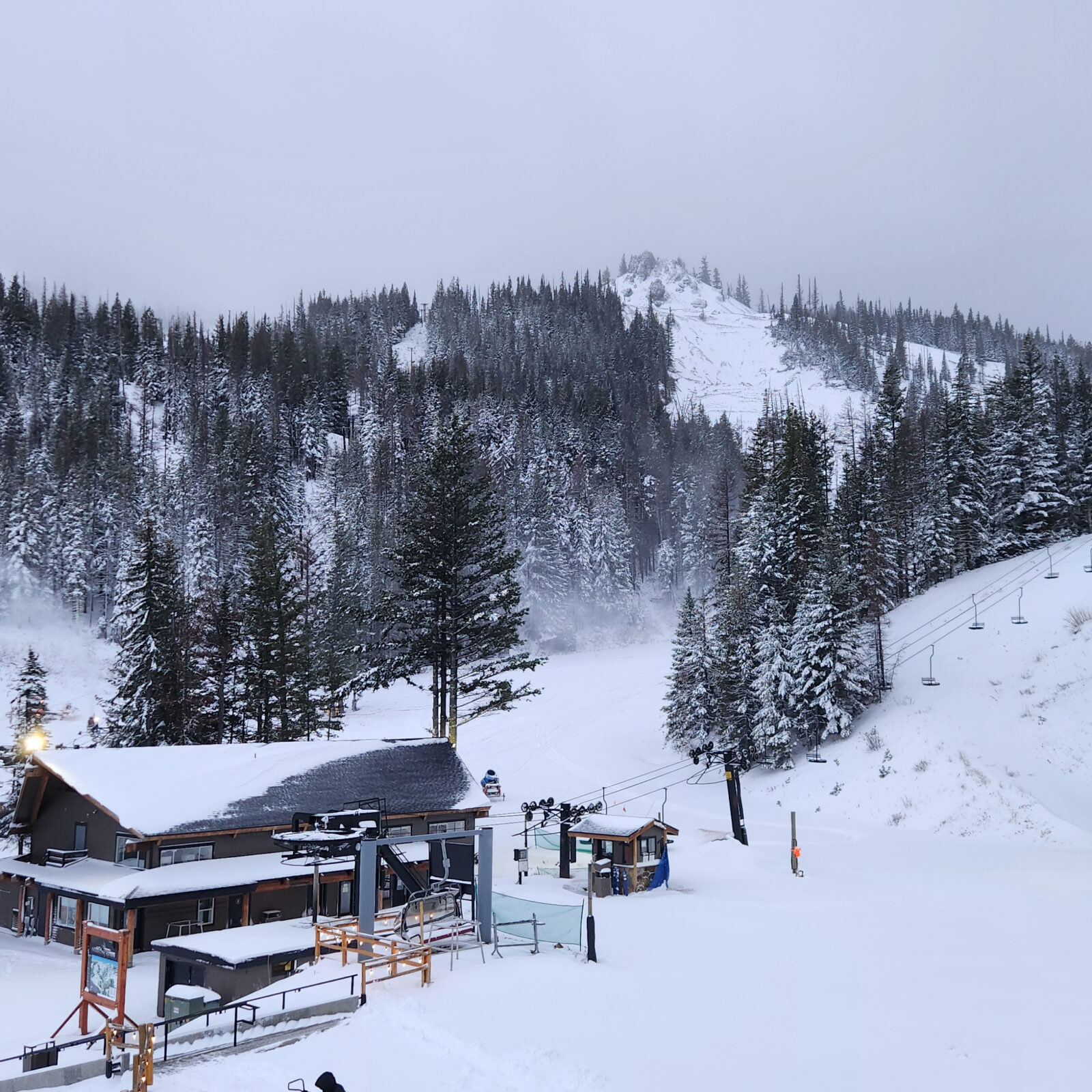 A snowy mountain scene at Mission Ridge showing the base area with a chairlift, lodge buildings, and snow-covered trees. Fresh snow blankets the ground and surrounding forest, with light snowmaking mist visible near the lower slopes. A ridge line rises in the background under overcast skies, creating a wintry, early-season atmosphere.