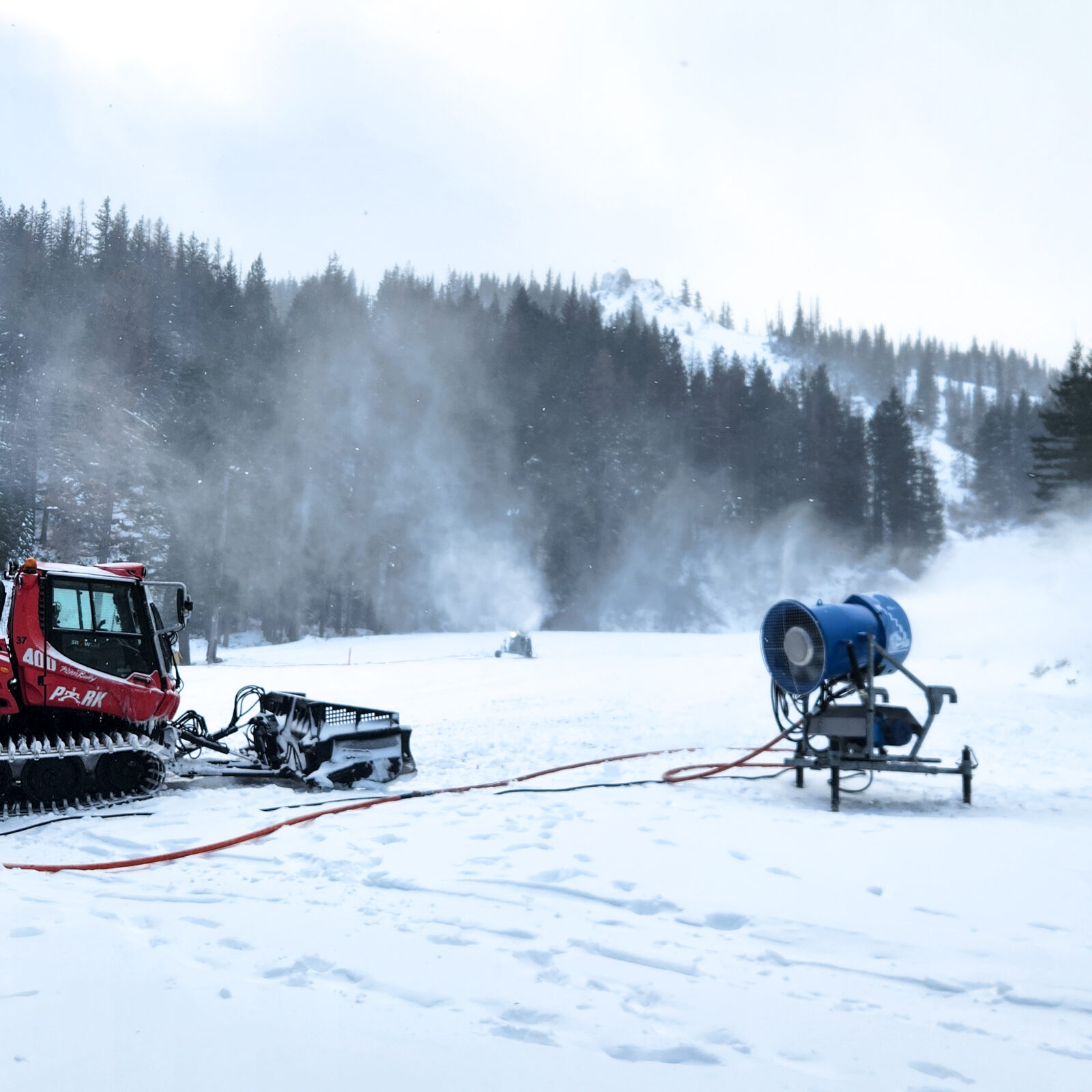 A red PistenBully snowcat and a blue snowmaking fan sit on a snowy slope at Mission Ridge as snowmaking mist rises across the run. Fresh snow covers the ground and the surrounding forested hillside, with low clouds and winter light creating a cold, overcast atmosphere. Orange snowmaking hoses stretch across the foreground.