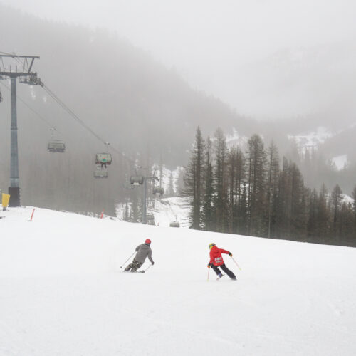 A young skier and father descend the Tumwater run at Mission Ridge on a foggy day, with Chair 2 and some evergreen trees in the background.