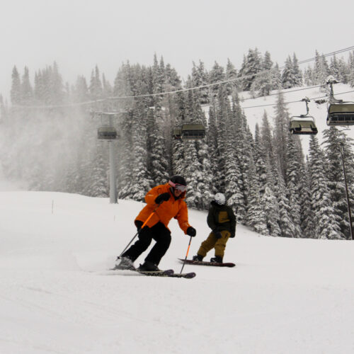 A skier and boarder ride together on a snowy, overcast slope