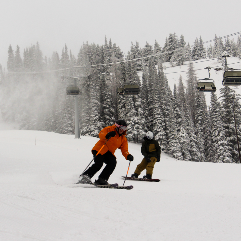 A skier and boarder ride together on a snowy, overcast slope