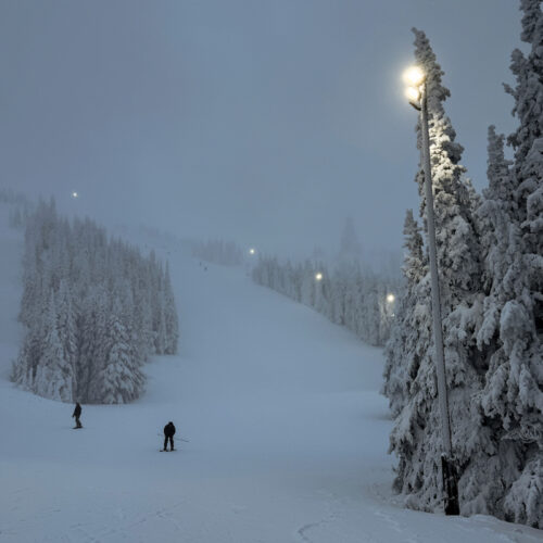 A view of Sunspot at Mission Ridge at night, with snowy trees, the night skiing lights aglow, and a skier and a snowboarder descending the slope.