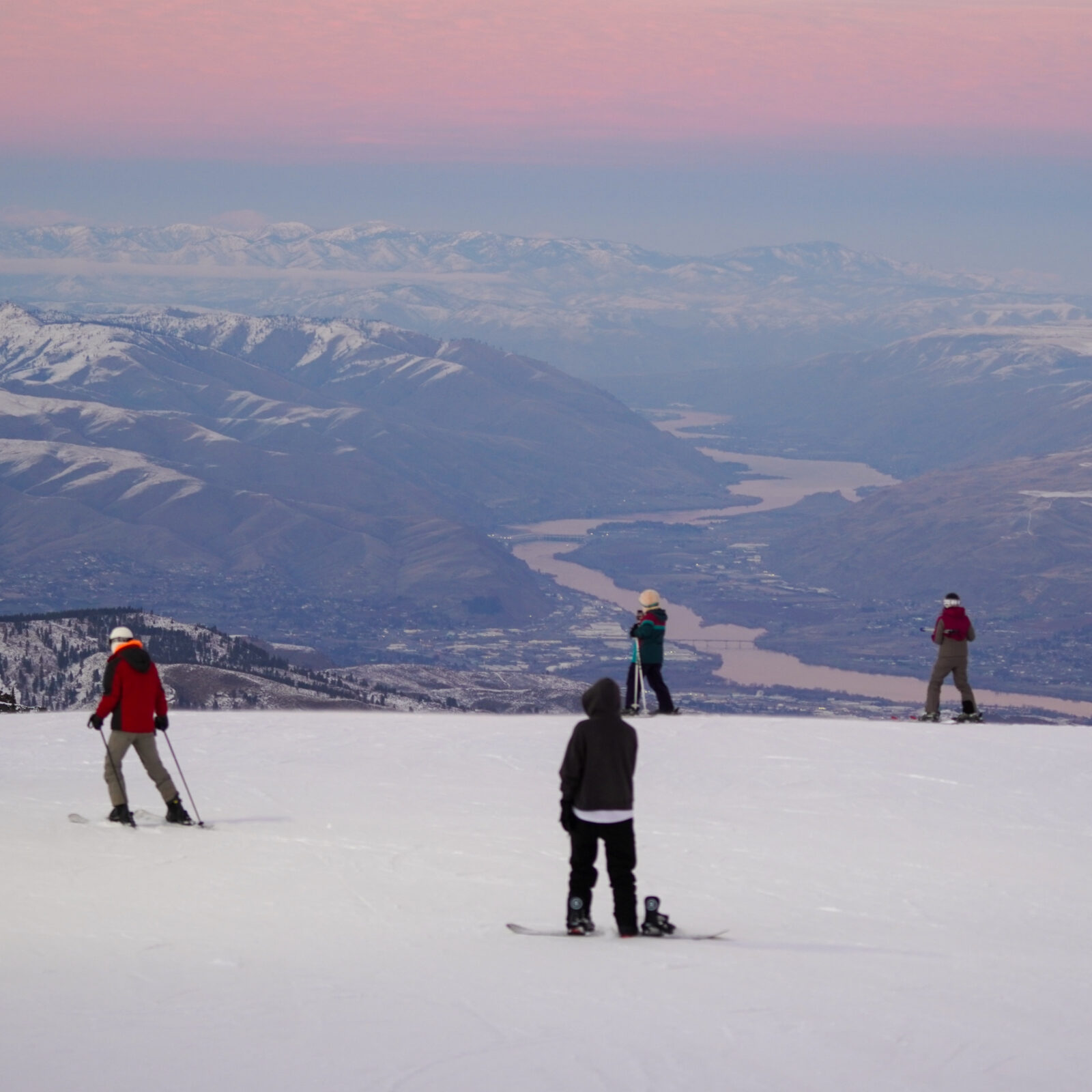 Skiers and snowboarders pause on a snowy ridge at Mission Ridge, looking out over the Wenatchee Valley and winding Columbia River below, with layered mountain ranges stretching into the distance under a soft pink and blue winter sky.