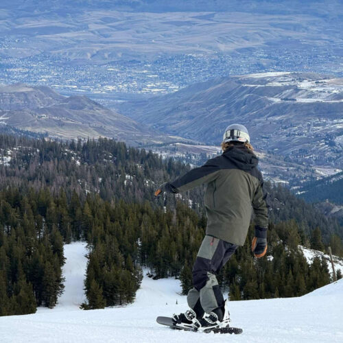 Snowboarder in a helmet rides down a snowy slope above a dense evergreen forest, with a wide valley and distant town visible under partly clearing skies.
