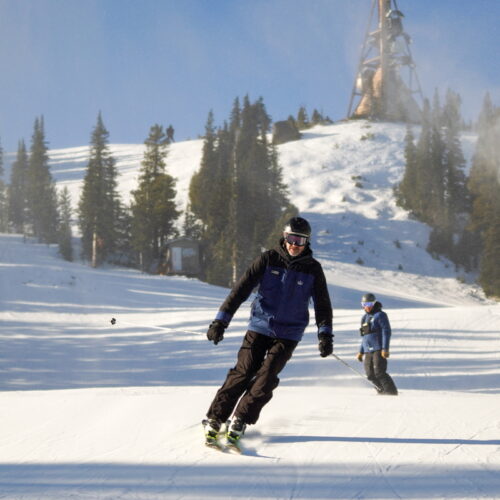 Skier dressed in dark blue skiing down sunlit slope, snowboarder in background.