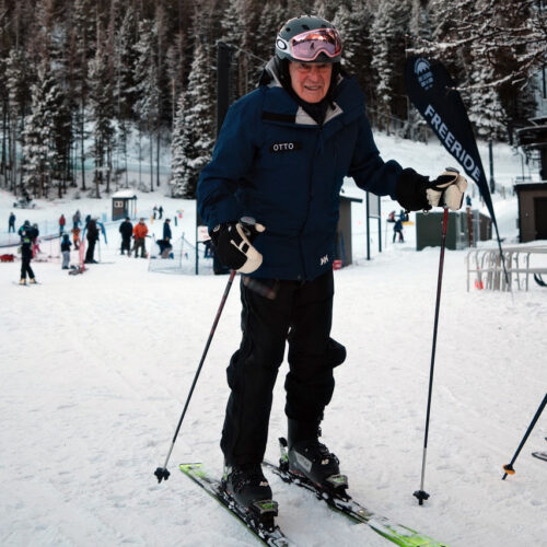 Otto Ross wearing a helmet and pink goggles smiles at the camera while standing on skis with poles at a snowy base area; a ‘Freeride’ flag and other skiers are visible in the background at Mission RIdge.