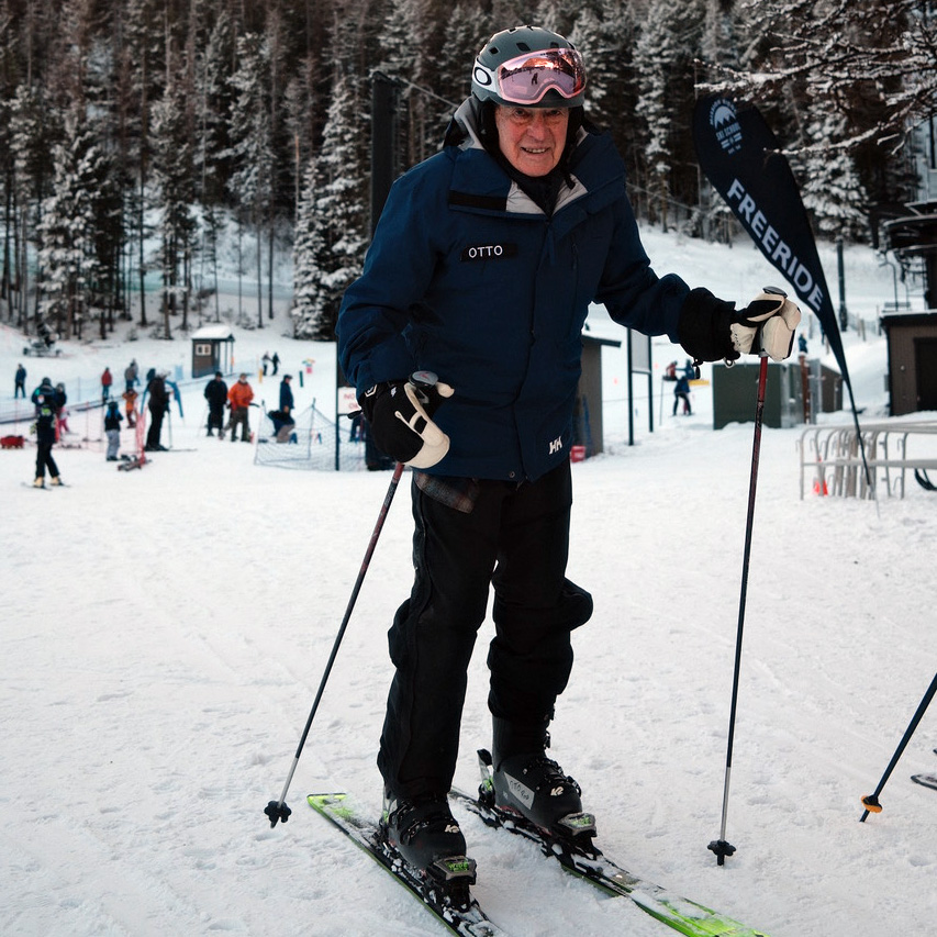 Otto Ross wearing a helmet and pink goggles smiles at the camera while standing on skis with poles at a snowy base area; a ‘Freeride’ flag and other skiers are visible in the background at Mission RIdge.