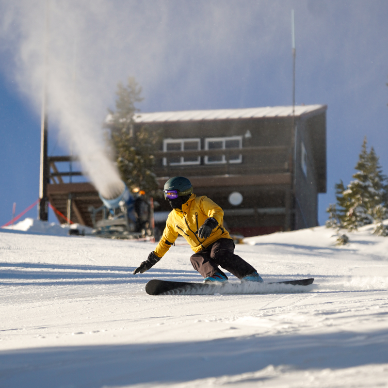 A skwal skier, with feet in line carves a sunny slope with a cabin and a running snow gun in the back ground