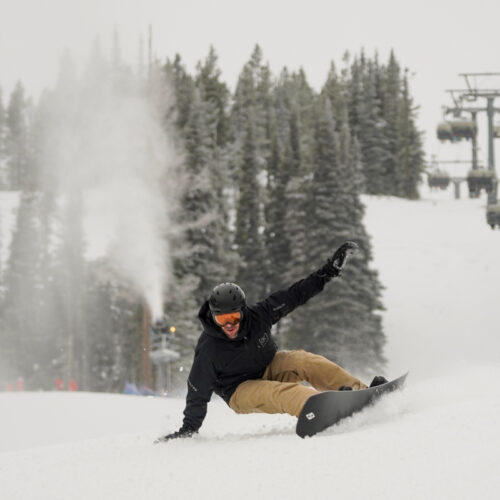 a snowboarder carves hard and drag his hand in the snow. a snowgun showers the lift line and trees in a white spray
