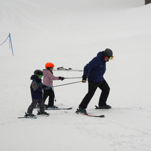 A ski instructor hauls two children with his ski poles on a flat section of a snowy run