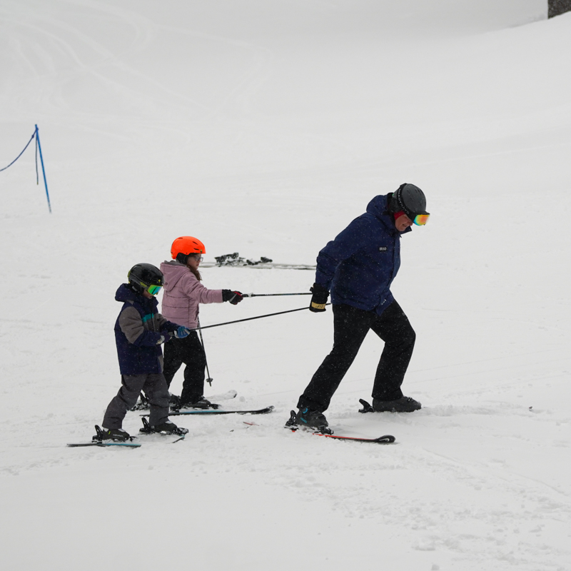 A ski instructor hauls two children with his ski poles on a flat section of a snowy run