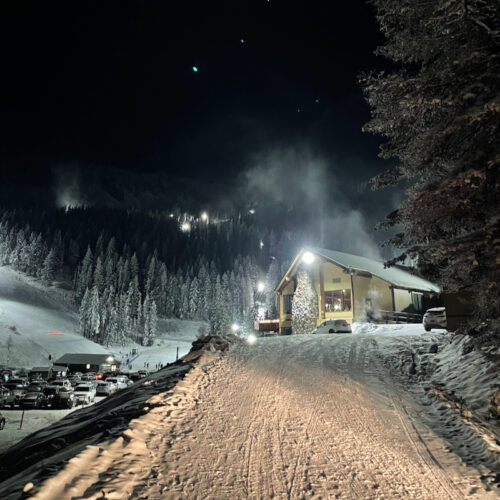 the Mission Ridge base area at night, showing dark snowy trees, cars in a snowy parking lot, and the lodge building on the right.