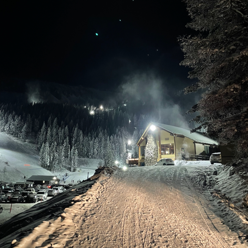the Mission Ridge base area at night, showing dark snowy trees, cars in a snowy parking lot, and the lodge building on the right.