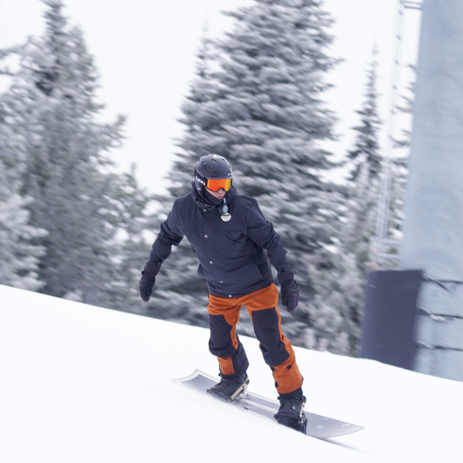 Snowboarder in orange pants slides past pole.