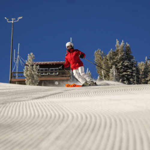 A skier descends the Sunspot Trail at Mission Ridge on a sunny day, with fresh corduroy in front and blue skies behind.