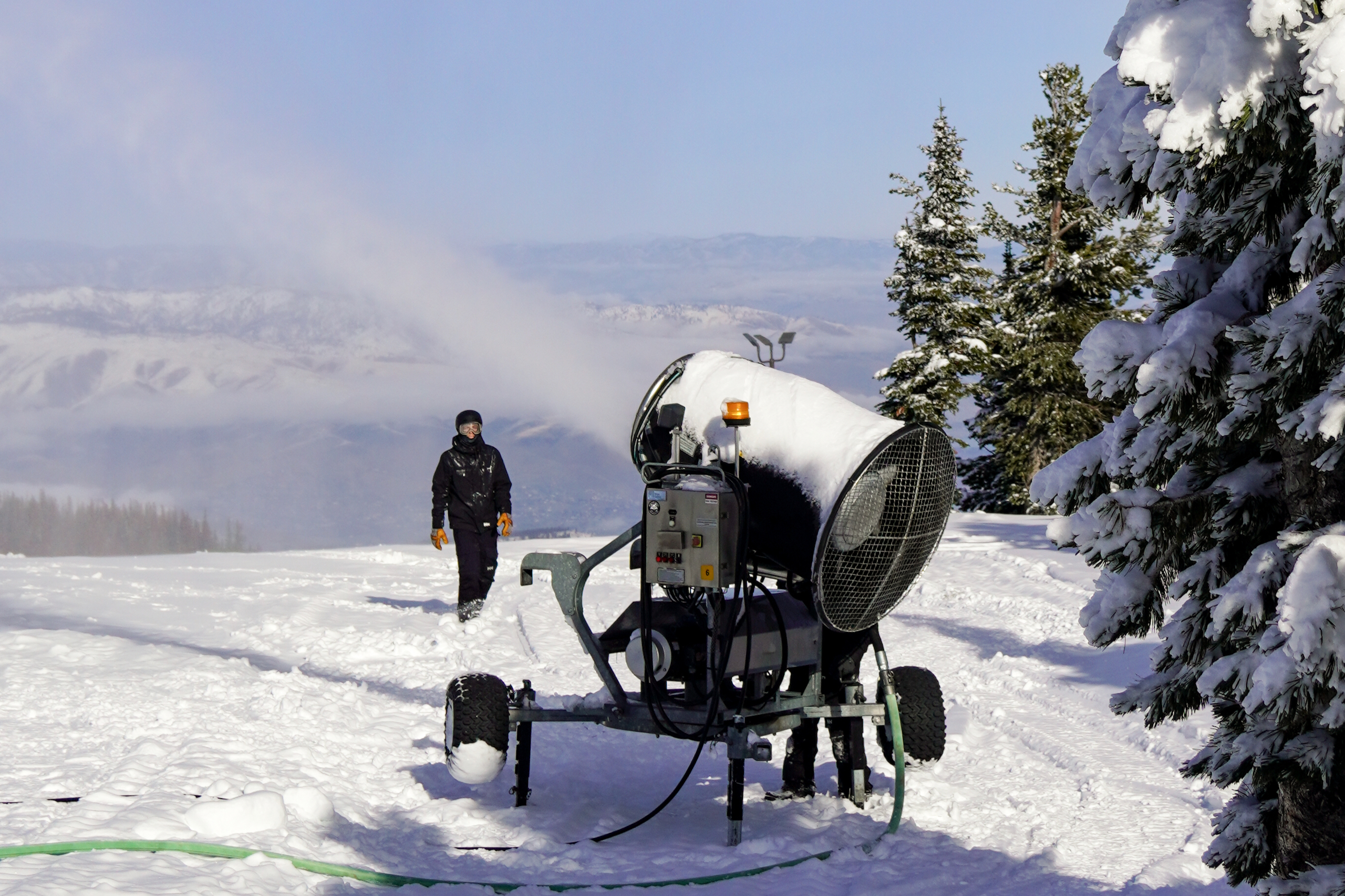 A snowmaker walks toward a snow gun firing near the top of Mission Ridge on a sunny day.