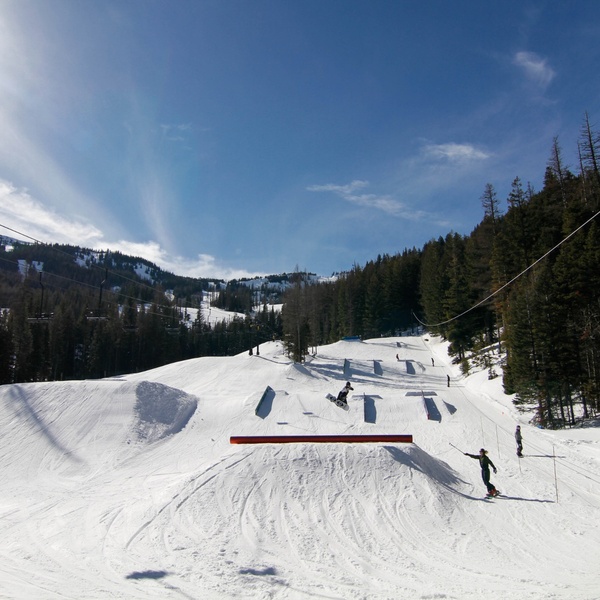 Snowboarder jumps over a red rail in a snowy terrain park, with ski lift lines, evergreen trees, and a bright blue sky in the background.