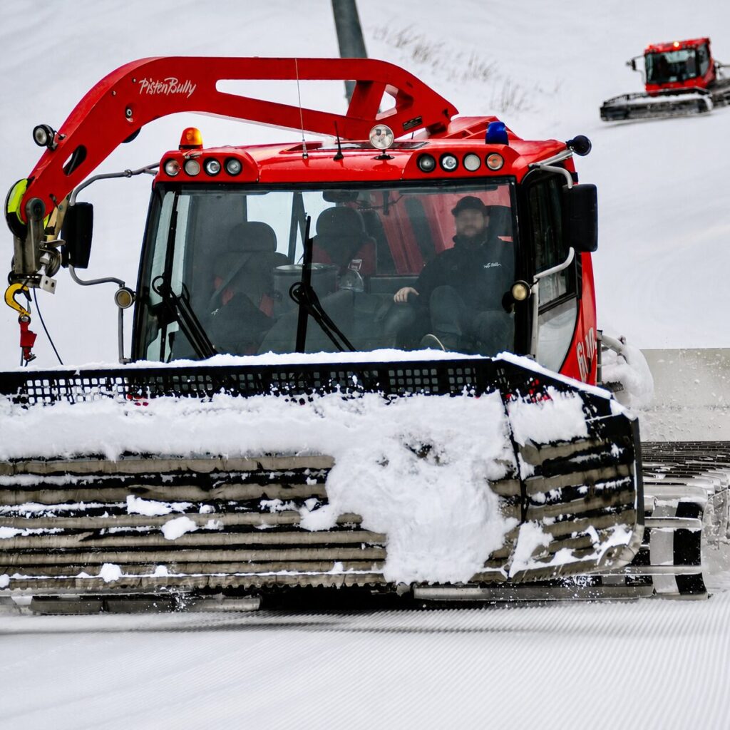 Close-up of a red snow groomer plowing a snowy ski slope, with an operator visible inside the cab and fresh corduroy snow in the foreground.