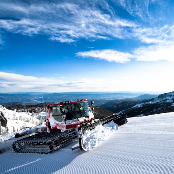Red snow groomer plowing a freshly groomed ski slope with snowy mountains, evergreen trees, and a bright blue sky in the background.