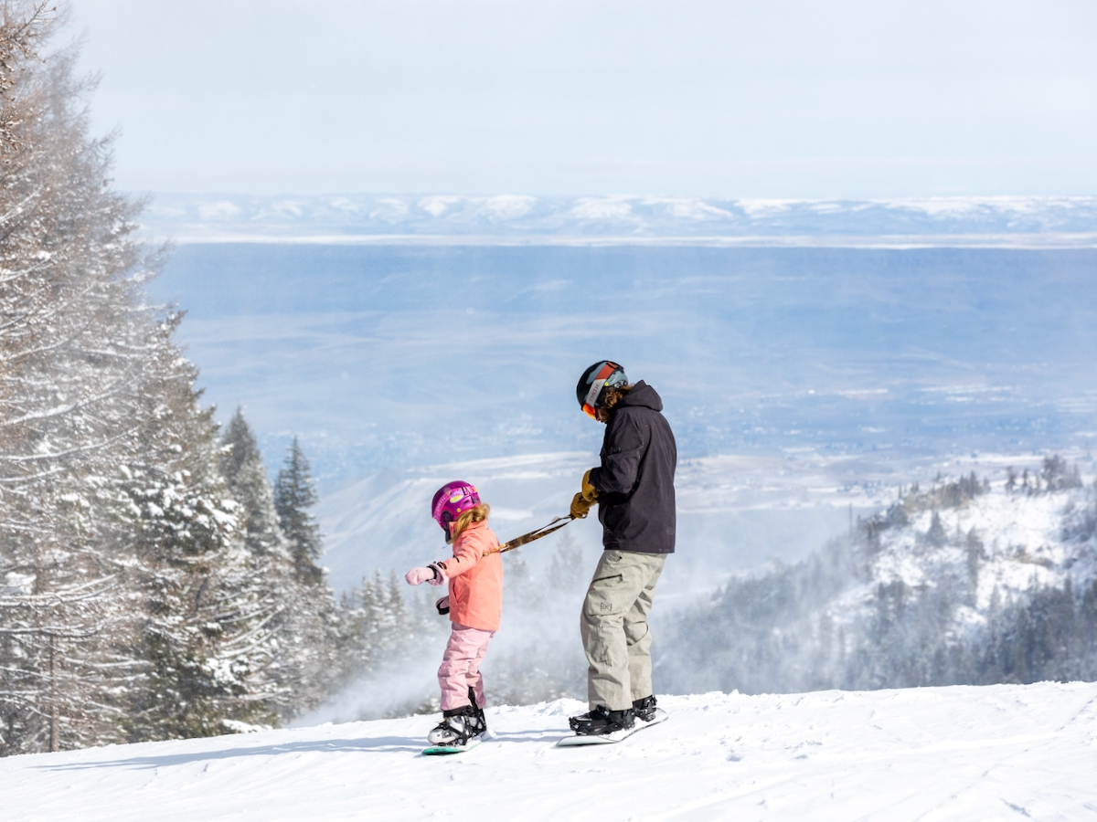 A parent helps a young snowboarder down a green run at Misison Ridge, with a view into the Columbia Valley below.