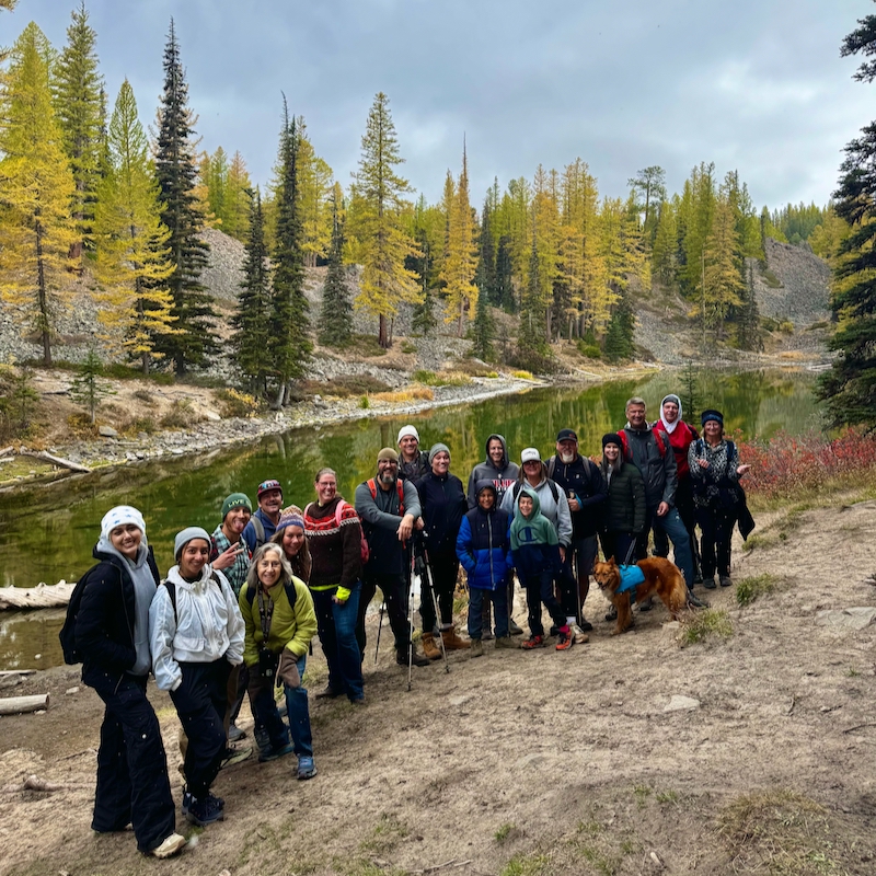 A group of hikers in front of Lake Clara in the fall, with golden larch trees lining the lake.