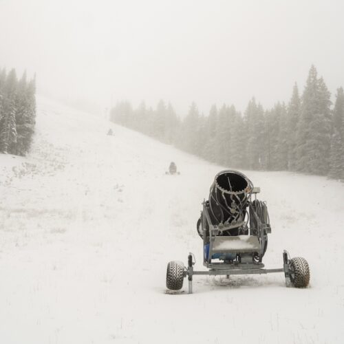 A snowmaking gun on a fresh coat of snow in the fog, with more guns lined up the slope and snowy evergreens on both sides