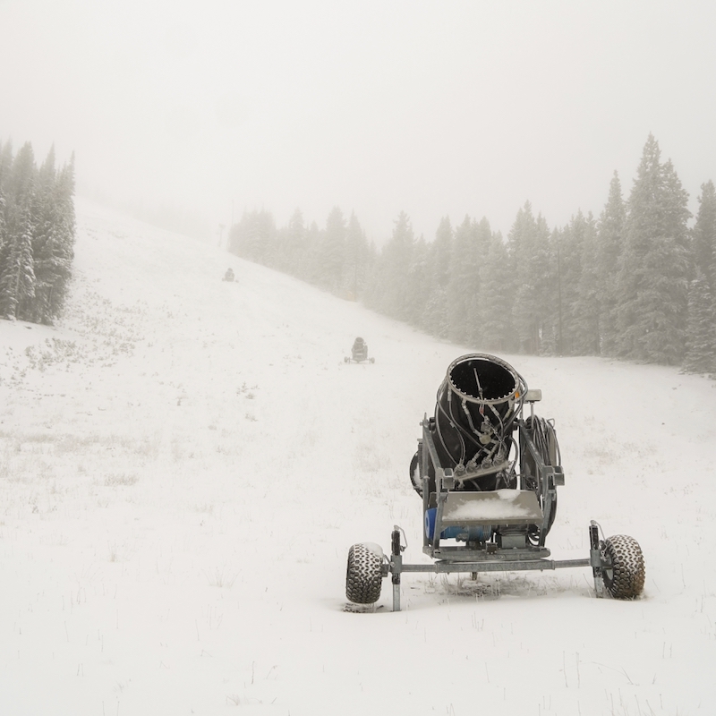 A snowmaking gun on a fresh coat of snow in the fog, with more guns lined up the slope and snowy evergreens on both sides